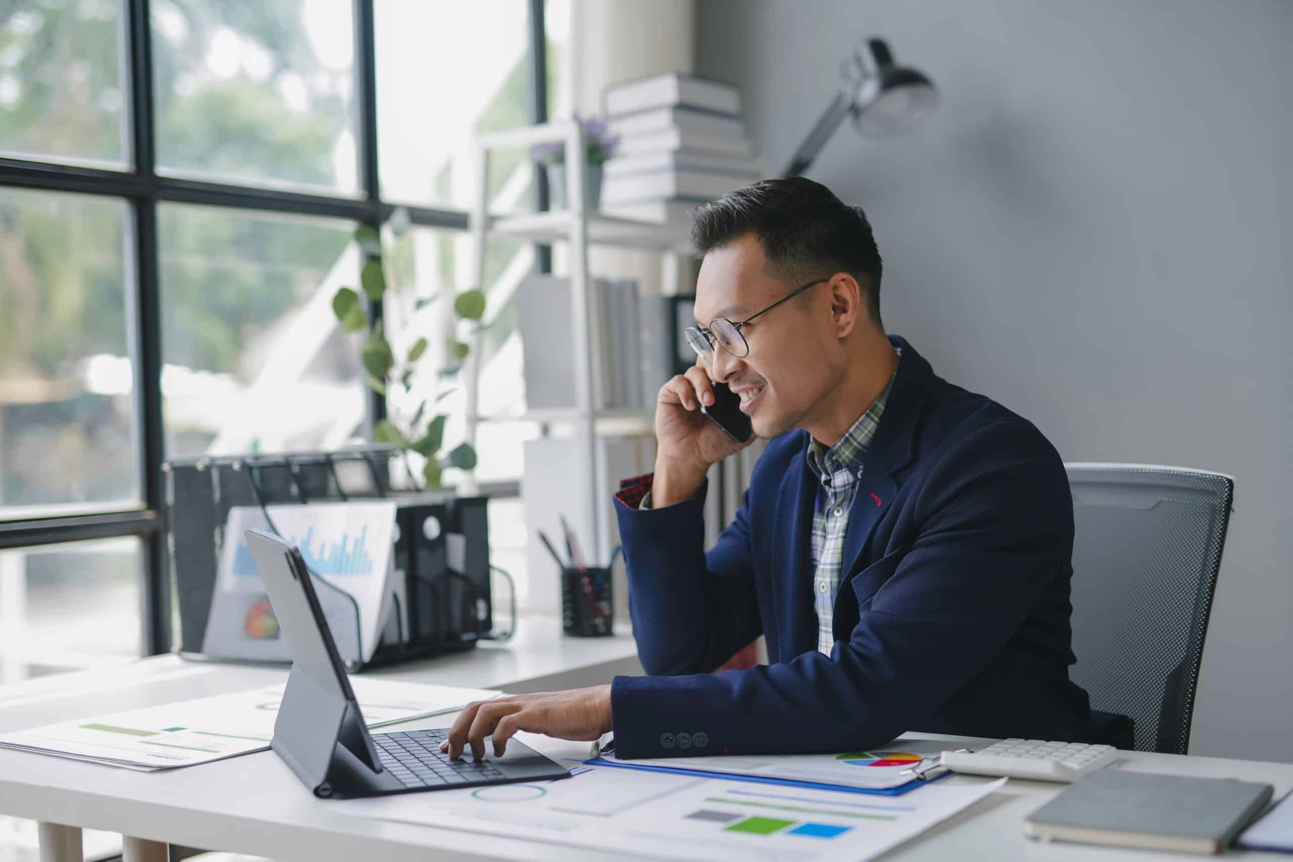 Businessman is smiling while working on a tablet and talking on the phone in his bright office